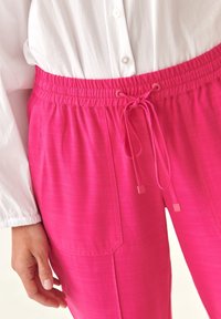 Růžové, lehké kalhoty s elastickým pasem, stahovací šňůrkou a bočními kapsami. Tkanina má jemnou texturu.