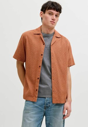 Młody mężczyzna w rdzawym, wzorzystym, krótkim rękawie koszuli nałożonej na szarą koszulkę z nadrukiem i jasnoniebieskich dżinsach, stojący na jednolitym tle.