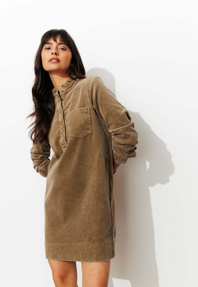 Robe en velours côtelé marron avec un bouton devant, un col et une poche poitrine unique. Manches longues avec poignets retroussés ; texture douce, longueur jusqu'au genou.