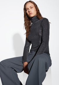 Top nero attillato con collo alto e una leggera lucentezza, abbinato a pantaloni grigi a gamba larga. Modella seduta su uno sfondo neutro.