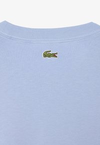 Camiseta azul claro de cuello redondo con un pequeño logo de cocodrilo verde bordado centrado debajo del cuello en la parte trasera.