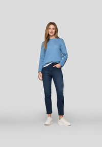 Eine Frau trägt einen blauen gestrickten Pullover, dunkelblaue Jeans und weiße Sneaker und steht vor einem grauen Hintergrund.