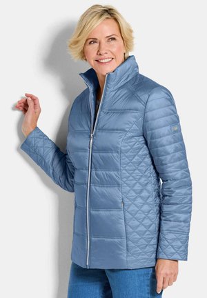 Lächelnde Frau trägt hellblaue gesteppte Jacke mit Stehkragen, posiert mit einer Hand an der Wand, leicht nach rechts gewandt.