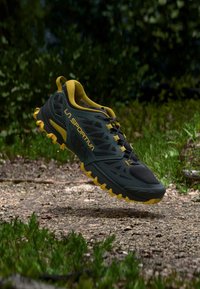 Črno in rumeno tekaško copato za steze z teksturiranim zgornjim delom, ojačanimi stranicami in agresivnim podplatom, zasnovano za zunanjo uporabo.