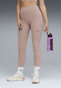 Leggings taille haute rose clair avec une texture mate et un petit logo Puma marron. Portés avec des baskets blanches et une bouteille d'eau rose.