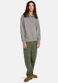 Felpa grigia con tasca a marsupio, abbinata a pantaloni larghi verde oliva con cuciture visibili. Scarpe marroni slip-on completano il look.