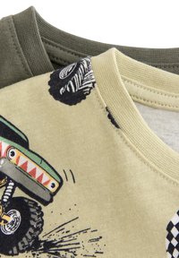 Dve zložené tričká s okrúhlym výstrihom, jedno olivovo zelené a jedno béžové s potlačou monster trucku a pneumatikových stôp na prednej strane.