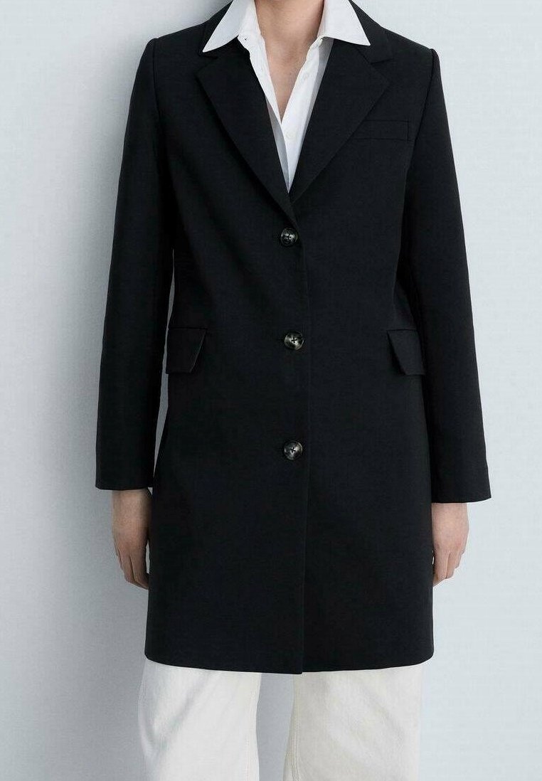 Personne portant un long blazer noir à trois boutons par-dessus une chemise blanche et un pantalon blanc, sur un fond clair uni.