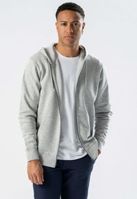 Gr å zip-up hoodie i blødt stof med foret hætte, snoretræk og frontlommer, båret over en hvid T-shirt og parret med mørke bukser.