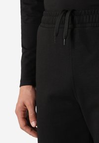Pantaloni della tuta neri con un tessuto strutturato, vita elastica e cordoncino con estremità, vista ravvicinata che mostra una mano che tiene il tessuto.