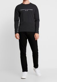 Schwarzes Langarm-T-Shirt mit weißem "TOMMY HILFIGER EST. 1985" Logo, kombiniert mit schwarzen Jeans und weißen Turnschuhen. Mit Rundhalsausschnitt.