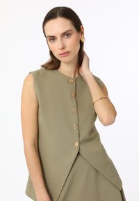 Blouse sans manches olive avec des accents de boutons dorés et un design ajusté. Texture lisse et détail d'ourlet asymétrique visible.