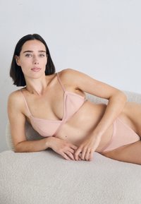 Conjunto de bikini em rosa claro, com detalhes em nervuras, alças finas e bordas em formato ondulado, apresentando um top triangular e uma parte de baixo de cintura baixa a condizer, sobre uma superfície branca texturizada.