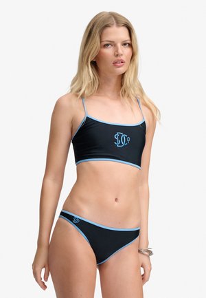 Blondynka w czarnym bikini z jasnoniebieskimi lamówkami i logo, stojąca z neutralnym wyrazem twarzy na białym tle.