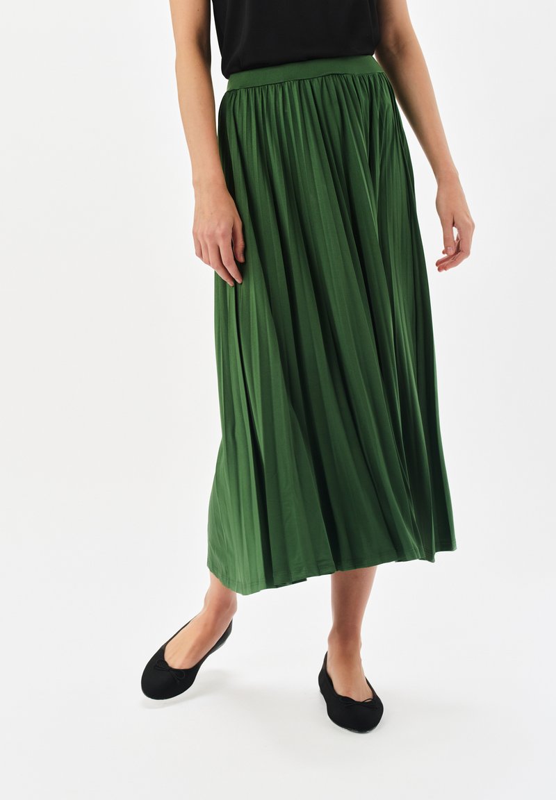 Zielona, plisowana spódnica midi z elastycznym pasem, wykonana z gładkiego, lekkiego materiału, zestawiona z czarnymi płaskimi butami na białym tle.
