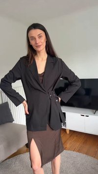 Γυναίκα με μακριά σκούρα μαλλιά που φοράει μαύρο σακάκι με ζώνη πάνω από καφέ φόρεμα με σκίσιμο στο μπούτι, στέκεται μέσα με τα χέρια στη μέση.