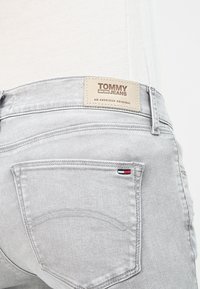 Vaqueros de mezclilla gris claro con un pequeño logo rojo, blanco y azul en el bolsillo trasero y una etiqueta beige de Tommy Jeans en la cintura.
