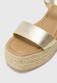 Sandali con fascia metallica dorata, dotati di una soletta imbottita color cammello e una suola piatta in juta intrecciata e testurizzata. Presentano una finitura liscia e lucida con dettagli di cucitura.