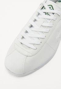Fehér bőr sneaker, texturált orr-résszel, fehér fűzőkkel, zöld részletekkel a nyelvén és diszkrét varrással az oldalon.