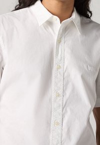 Weißes Hemd aus leichtem Stoff. Verfügt über einen klassischen Kragen, sechs Knöpfe vorne und eine dezente Struktur.