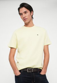 Νεαρός άνδρας με σκούρα μαλλιά, φορώντας ένα ανοιχτό κίτρινο t-shirt Calvin Klein, σκούρα τζιν και μαύρη ζώνη, που στέκεται με τα χέρια στις τσέπες.