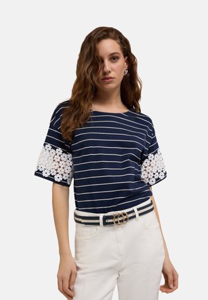 Marineblauw en witte gestreepte top met korte mouwen en bloemenkantaccenten op de mouwen. Gecombineerd met witte broeken en een tweekleurige riem met een ronde gesp.