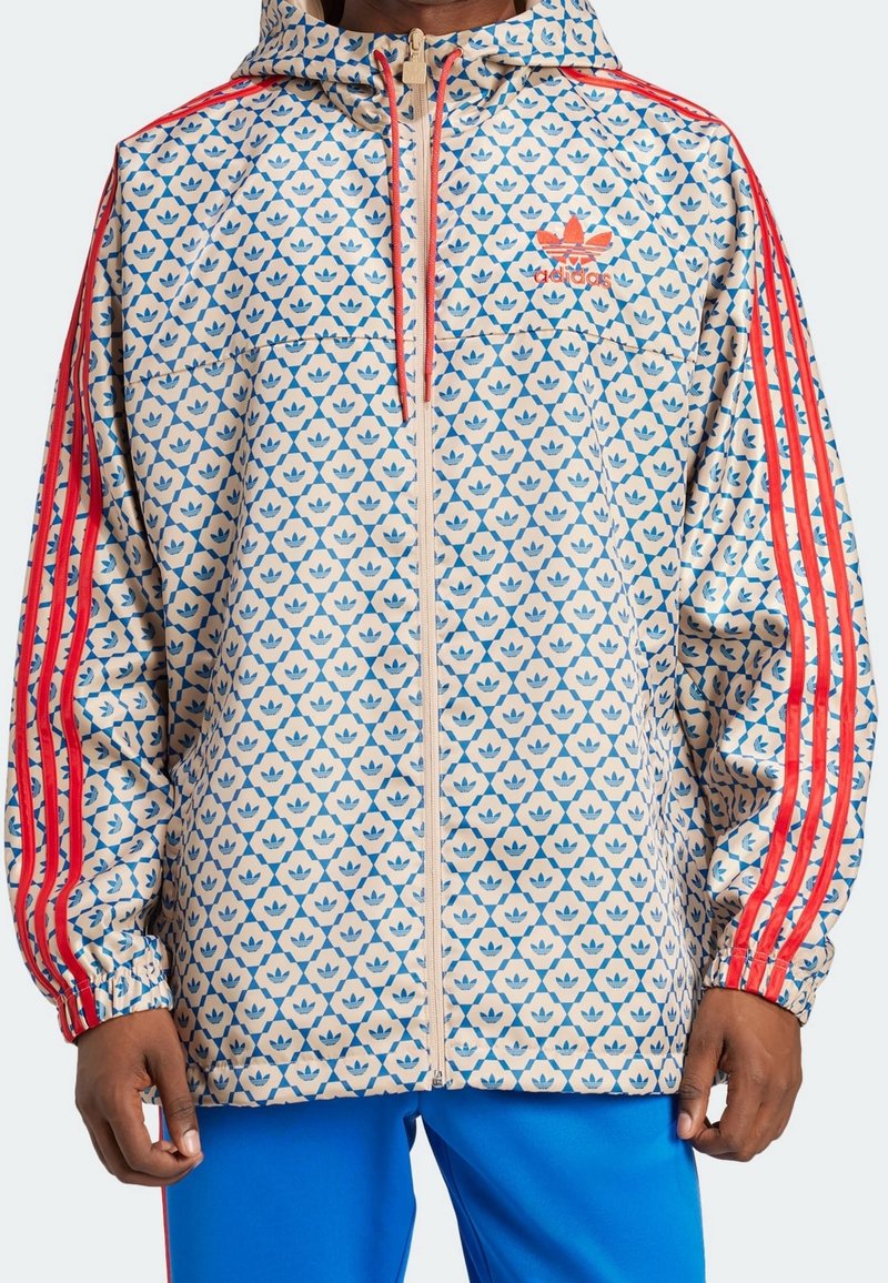 Chaqueta beige con patrón de hojas azules, detalles en franjas rojas en las mangas, cierre frontal de cremallera completa, puños elásticos y capucha. Material suave y ligero.