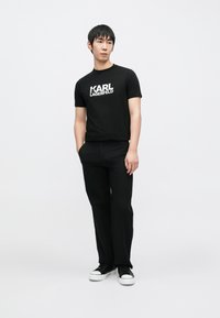 Fekete pamut póló fehér "KARL LAGERFELD" logóval, fekete nadrággal és fehér talpú fekete sportcipővel párosítva. A modell neutrálisan áll.