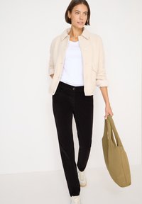 Beige gestructureerde jas, witte T-shirt, zwarte corduroy broek en lichte sneakers. Houdt een groene tas vast. Eenvoudige, casual outfit.