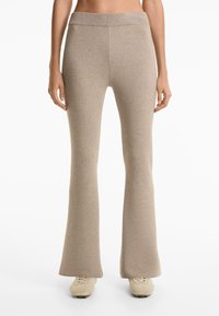Vrouw in beige high-waisted gebreide flare pants en beige sneakers, staande voor een effen witte achtergrond.