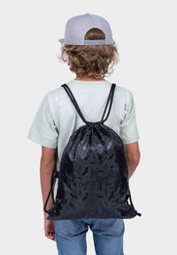 Black drawstring backpack with bat pattern; made of lightweight fabric. User wearing a mint-green shirt and jeans, seen from behind.
