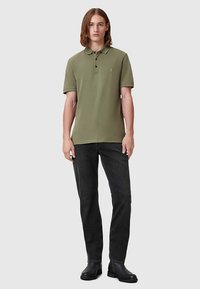 Olívazöld galléros póló három gombbal, sötétszürke farmernadrággal és fekete bokacsizmával párosítva. Sima szövet textúra, egyszerű kialakítás.