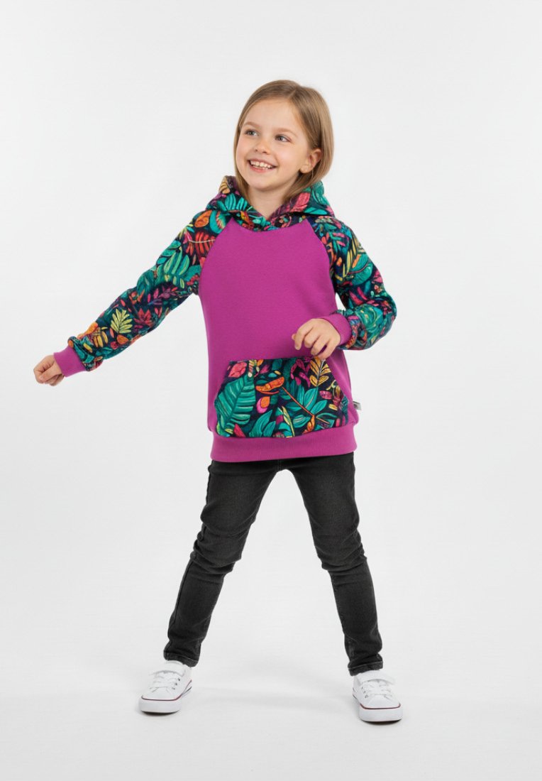 Uśmiechnięte dziecko w fioletowej bluzie z kapturem ozdobionej kolorowymi wzorami liści oraz w czarnych spodniach, stojące z rozstawionymi nogami i lekko podniesionymi ramionami.