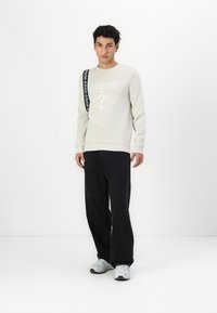 Sudadera gris claro con el logo "GUESS", combinada con pantalones negros y zapatillas grises. Presenta una correa negra con detalles del logo en el hombro.
