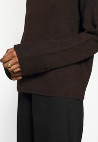 Ruskeat neulotut villapaidat, joissa on tekstuuri, leveillä resoreilla ja rento malli, yhdistetty mustiin housuihin. Käsi on koristeltu kultaisella sormuksella.