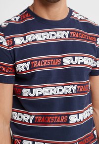 Mørkeblå bomulds t-shirt med vandrette striber og teksten "SUPERDRY" og "TRACKSTARS" i hvid og rød. Rund hals og korte ærmer.