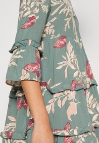 Robe fleurie en tissu vert avec des manches en cloche. Présente un design superposé, des feuilles roses et des papillons dans des teintes de bourgogne et de crème.