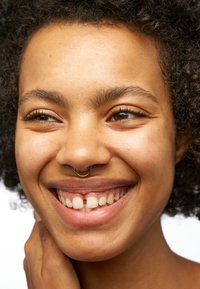 Gros plan sur une personne avec des cheveux bouclés et foncés, portant un anneau septum en or. Peau lisse, maquillage naturel et un large sourire avec des dents visibles.