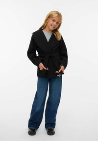 Cappotto nero avvolgente con ampio colletto, vita cin cin, e vestibilità morbida, abbinato a jeans a gamba larga blu e scarpe nere. Tessuto dalla trama liscia.