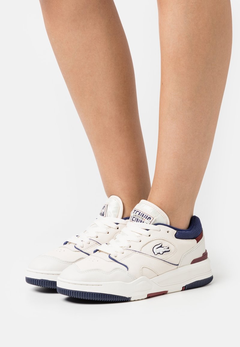Baskets blanches Lacoste avec des touches bleu marine et bordeaux portées aux pieds sur un fond blanc uni.