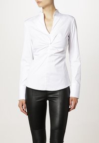 Camicia bianca a maniche lunghe con dettaglio avvolgente sul fronte, caratterizzata da un colletto a punta e una texture liscia e croccante; abbinata a pantaloni neri in pelle aderenti.