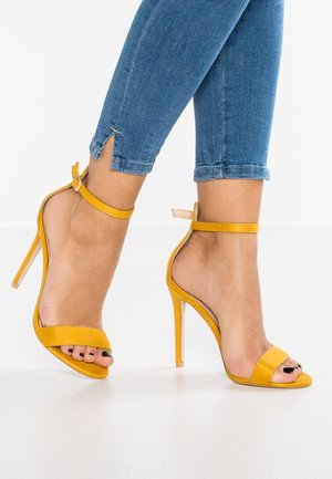 Żółte zamszowe sandały na wysokim obcasie z pojedynczym paskiem na palce i paskiem na kostkę. Cechują się smukłym obcasem typu stiletto oraz otwartym tyłem.