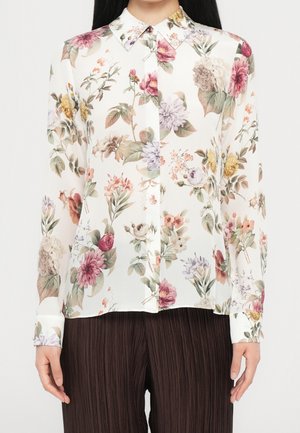 Weiße langärmelige Bluse mit Knöpfen und mehrfarbigem Blumenmuster, getragen zu dunkler plissierter Hose.