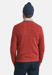 Rot gestrickter Pullover mit Rundhalsausschnitt und gerippten Bündchen, kombiniert mit einer dunkelblauen gerippten Mütze und einem blauen Hemd darunter. Dunkle Jeans sind sichtbar.