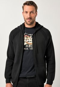 Schwarzer Zip-Hoodie mit texturiertem Finish, ausgestattet mit einer Kapuze mit Kordelzug und zwei Fronttaschen, über einem schwarzen Grafik-T-Shirt.