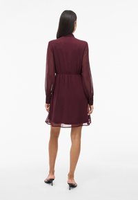 Kobieta z długimi ciemnymi włosami, ubrana w burgundową długą sukienkę z przezroczystego materiału i czarne szpilki, stojąca tyłem.