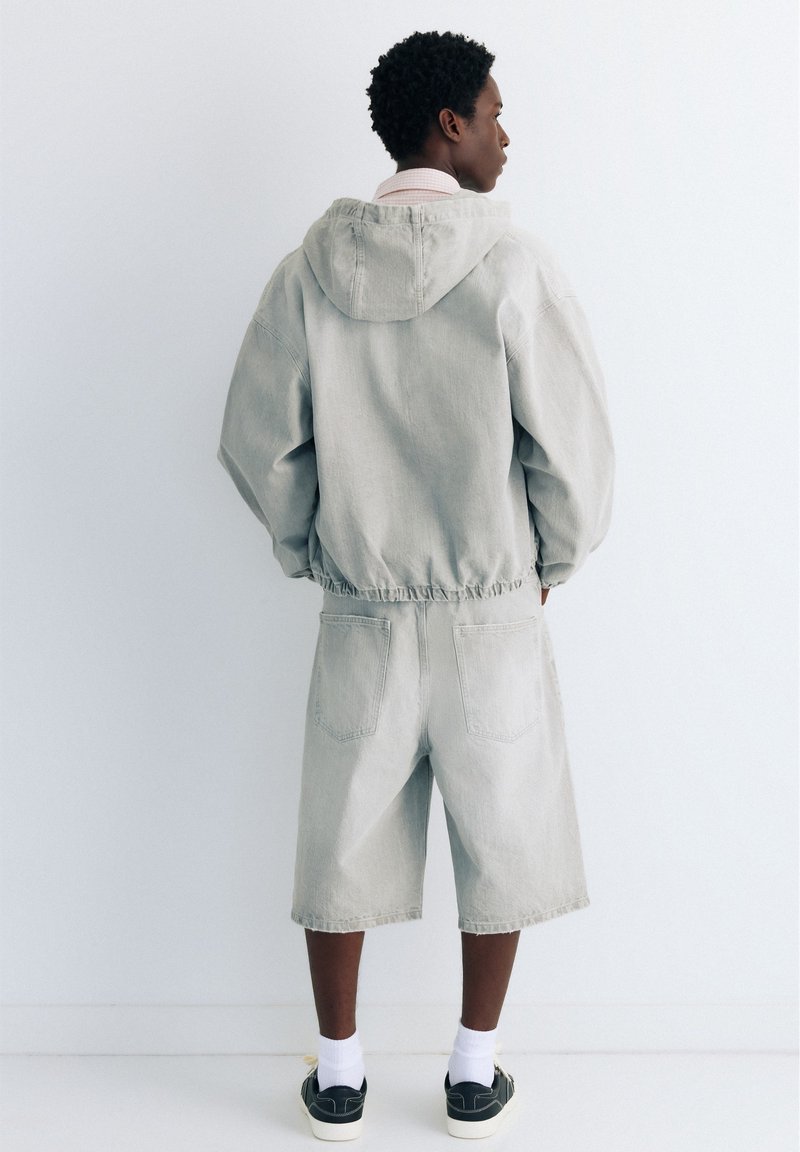 Jeune personne debout face à un mur, portant une veste à capuche gris clair, un short long assorti, des chaussettes blanches et des baskets noires.