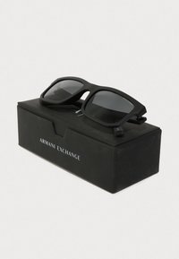 Melnas saulesbrilles ar taisnstūra lēcām matēta apdare, atpūšoties uz melnas kastes ar uzrakstu "Armani Exchange." Kastei ir vienkārša dizaina.