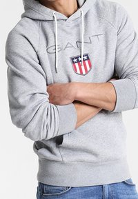 Person in einem hellgrauen GANT-Hoodie mit weißen Kordeln, die Arme verschränkt, und in blauen Jeans, gezeigt von Hals bis Hüfte.