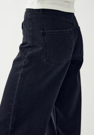 Close-up of person wearing black denim jeans showing back pocket and side seam against light background.